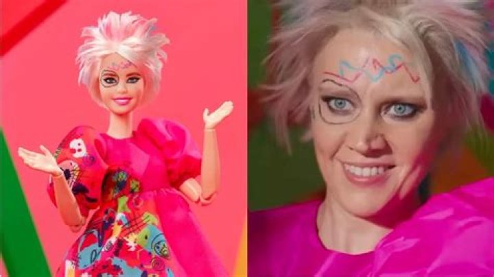 Kate McKinnon as Weird Barbie is holding up two different shoes.